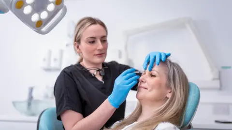 Picture of blonde haired lady smiling on a reclining seat in a aesthetic clinical setting. A lady in blue scrubs and blue gloves is about to inject near her right eye.
