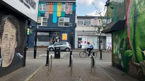 Handrails in a pedestrian area of Withington's high street with a road in the background. A cyclist cycles on the road, while flanking the pedestrian area are two murals, one featuring a face and the other some sort of green artwork. 