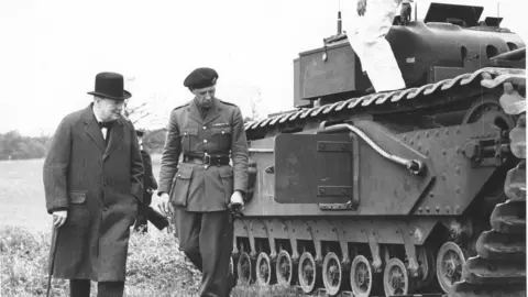 Prime Minister Winston Churchill in a coat and bowler hat inspecting a tank with a man in full uniform