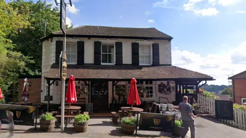 A two storey building painted white with black shutters and dark roof tiles. There are red umbrellas, picnic benches, silver kegs, and Guinness branded wind shields outside the property.