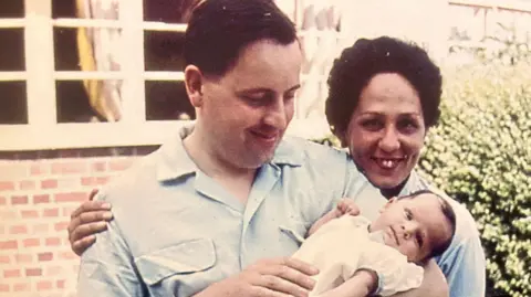 Chris Martin A mother and father stood for a photo. The woman is smiling at the camera outside a house with her arm around the father, who is looking down at a baby in his arms