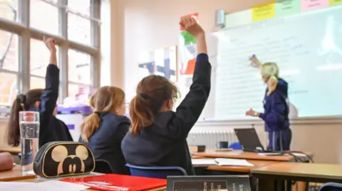 PA Female schoolchildren sat in a classroom, with a female teacher writing on the board. 