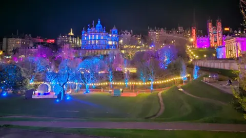 Nicola Campbell Un paisaje urbano nocturno con edificios históricos y árboles iluminados con luces de colores, creando una atmósfera festiva.