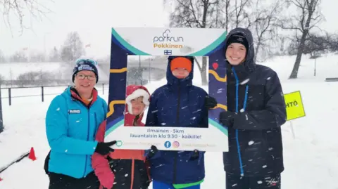 Clara Geraghty A family of runners are standing holding a cardboard picture frame which shows they are in Finland. It is snowing and there is white snow lying on the ground all around them.
