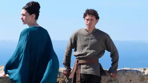 Tom Holland and Anne Hathaway in a film still from the film The Odyssey. Tom Holland is leaning against a wall with a scenic greek sea view behind him. He is wearing a dark olive green shirt and dark trousers. Anne hathaway is looking to the left and wearing a flowing flowing blue dress