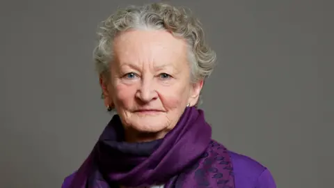 House of Lords A woman with short, wavy grey hair and wearing a purple scarf and purple jacket, stands in front of a dark grey wall.