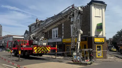 Fire appliance with aerial ladder parked outside a terrace of shops with flats above with its roof missing.