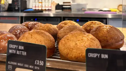 A selection of crispy buns on a cafe counter, both with butter and plain. A sign can be seen reading Crispy Bun with Butter, £3.40. A counter behind contains bottles of coffee syrup, food tubs and chopping boards.