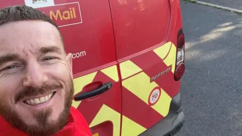 A picture of postman Adam Prowse stood behind his Royal Mail van. He has a short brown beard and is wearing a red jacket.