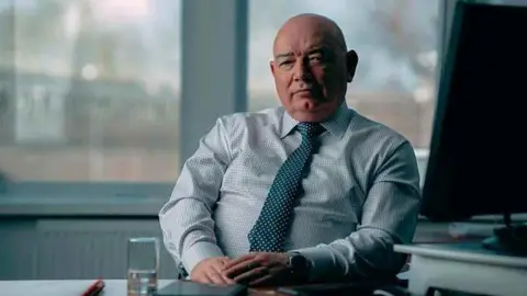 Mick Stoney sits behind a table in an office, wearing a plaid shirt and green tie