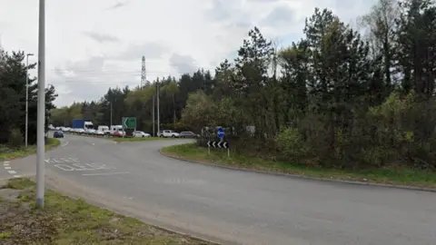 A large roundabout with trees in the centre. On the left of the picture is a long queue of cars waiting to join the roundabout.