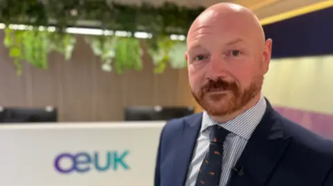 Man in a navy suit and patterned tie standing in a modern office reception area, with a blurred “OEUK” logo on the desk behind him and greenery hanging from the ceiling