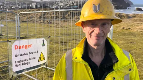Mining engineer Chris Rogers wearing a fluorescent yellow jacket and yellow hard hat, standing beside a fence around a hole that has opened up in sand dunes at Perranporth