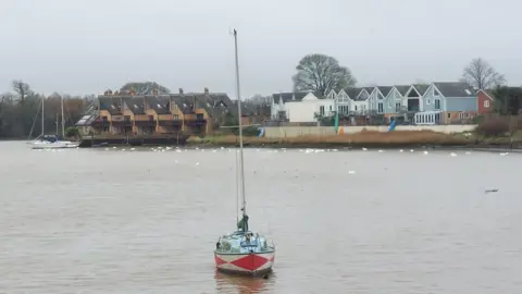 A sailing boat moored in a river with a light blue and red hull and a mast. On the far bank are two rows of houses. One is brick, the other is blue and white There are several white birds in the water. The sky overhead is overcast.