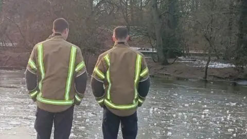 Two firefighters, pictured from behind, look across a frozen pond with trees in the distance