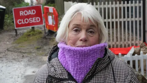 John Fairhall/BBC Maureen Jones wearing a black and white checked coat with a purple snood. She is looking directly into the camera. Behind her is a red Road Closed sign. 