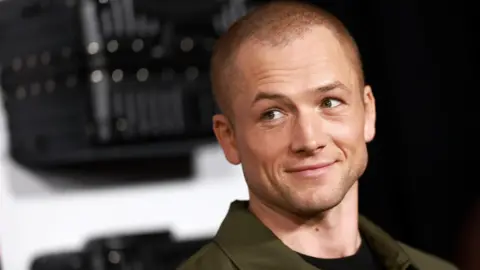 Getty Taron Egerton attends the premiere of Netflix's  Carry-On at The Egyptian Theatre Hollywood on December 5, 2024. He is wearing a khaki jacket and black t-shirt. He has a shaved head and is smiling to the left of the camera. 