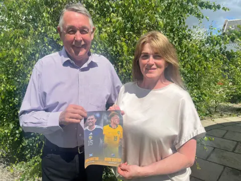 Souttar family Jack and Heather Souttar in blue and white shirts standing side by side smiling in front of a tree. They are holding photos of their sons in their international strips.