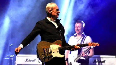 Francis Rossi wearing a black zip up jumper and a white shirt. He is standing on stage playing an electric guitar while another band member can be seen smiling and playing another white guitar behind him.