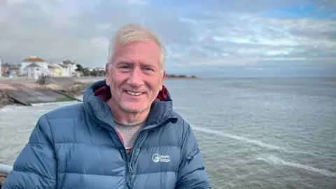 Derek Johnson wearing a mid-blue padded jacket with Exmouth seafront in the background.