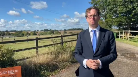 Ella Harget-Dash/BBC Alex Burghart is facing the camera wearing a navy suit with a light blue tie and white shirt. He has brown hair and is wearing glasses. He stands on a track road with fields behind him and electricity lines.