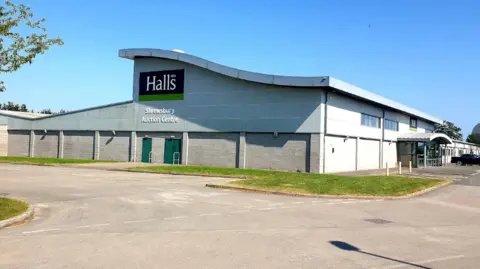 Halls A large grey building with a curved roof, a large car park at the front and a black sign which reads Halls