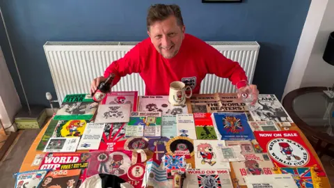 A man in a vintage red England shirt smiling in front of a table which is covered in 1966 World Cup memorabilia. 
