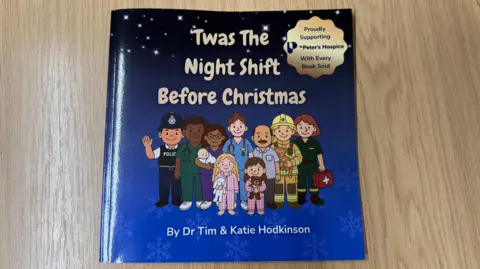 The front cover of a dark blue book with stars at the top of the cover. It shows a group of illustrated night-shift workers including doctors, nurses, firemen, paramedics and police standing in the middle. At the front of the group there are two small children wearing pyjamas and holding teddy bears.