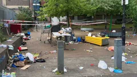 Side of a road with rubbish everywhere, including lots of bags and bottles. Police tape can be seen in the background. 