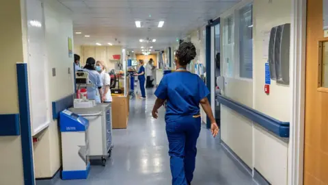 PA Media A nurse wears blue scrubs as she walks through a hospital corridor.