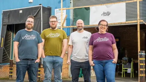 Amity Brew Co Three men and a woman wearing Amity-branded T-shirts stand in front of a brewery.