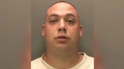 A police head shot of Joel who has a buzz cut hairstyle and is clean shaven. He is wearing a white t-shirt and is looking into the camera.
