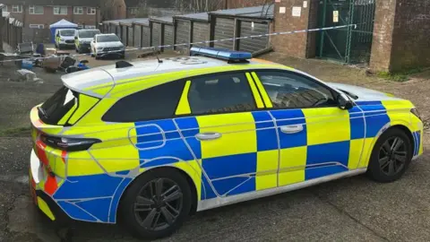 Kimberley Poper/BBC A police car, by police tape, by a row of garages and at the end of the row a police tent and three more vehicles. A pile of rubbish is outside a garage and there are houses at the end of the road. 