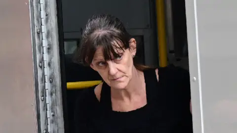 A woman with dark hair walking out of a police van. She had a black jumper dress on