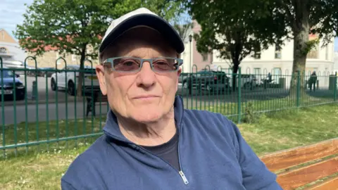 A head and shoulders photo of man wearing a white baseball hat and glasses, who is looking into the camera. He's sitting on a brown wooden bench and there is grass and a metal fence behind him, and some trees and cars beyond that. It's a bright day and the sun is shining on his face. He's wearing a navy blue jumper with a zip at the top, zipped down, with a black t-shirt underneath.