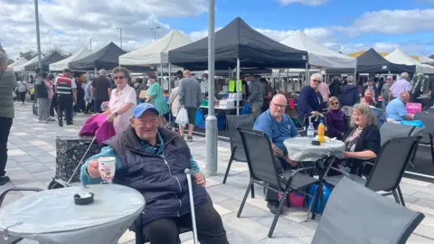 A bustling outdoor market with people sat on chairs at tables in the foreground. Stalls with canopies can be seen in the background with people looking around them.