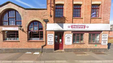 Google Two-storeys of the Phipps Brewery building in Northampton, which has windows with a slight arch to the top, separated into small red-framed sections. There is a white banner over the door with PHIPPS in red writing and various adverts for "ale and stout" attached to the brick walls. There is a pavement in front of the building.