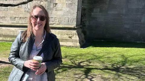 Carla Linford, a woman with long light brown hair, is standing in front of a large stone building. She is smiling for the camera while cupping a takeaway coffee cup.