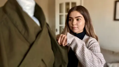 BBC/ Nicola Rees A young white woman with long brown hair and brown eyes makes adjustments to a khaki coloured coat on a mannequin 