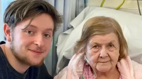 Tristan Essex A young man in a blue t-shirt is smiling and crouching next to an older woman in a hospital bed wearing a pink dressing gown. 