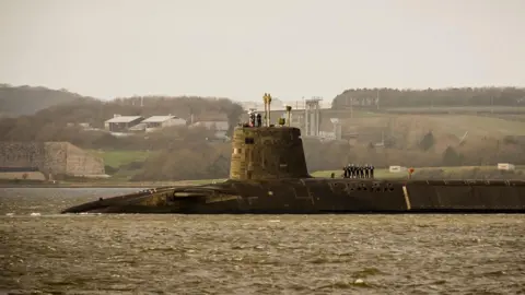 A submarine coming into port in Plymouth. There are officers standing in a line on the main body of it and a couple on the turret. It is a grey day and the sea looks brown. 