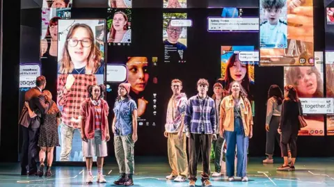 The cast of Dear Evan Hansen stand on the stage to sing You Will Be Found. Behind them are images of people appearing on screens on social media platforms. The six perfomers in the middle of the group are singing towards the audience. The other four characters are looking at the screens, two are standing upstage right and two upstage left.