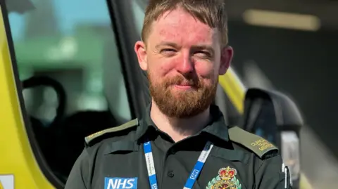 A paramedic wearing a dark green uniform which has an NHS label on. It also has an ambulance badge on it and he is also wearing a blue and white NHS lanyard. The man has auburn hair and and a beard and is smiling at the camera. 