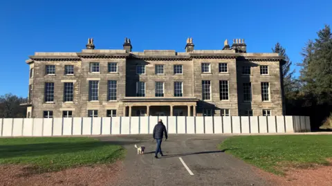 Haigh Hall in Wigan, a manor house surrounded by wooden boarding. There is a man with a dog in the foreground. The building is three storeys high, and features rows of large windows, with small chimney stacks seen across the flat roof. 