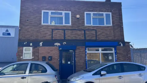 The two-storey building has blue paintwork around its entrance and lower window frames. There are two windows on the second floor and two cars are parked outside.
