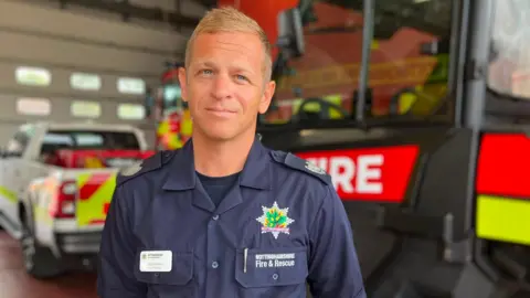 A man in a fire uniform in front of a fire truck. he has blue eyes and blonde hair 