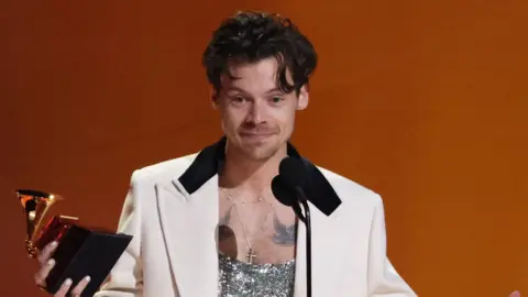 Reuters Harry Styles wears a white blazer with a black collar and is wearing a silver top. He is holding an award on stage and is stood in front of a mic