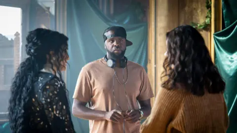 Daniel Alexander stood talking to two women who have their backs to the camera. Daniel Alexander is wearing a light pink t-shirt, ablack hat turned to the side and black Sony headphone around his nexk.