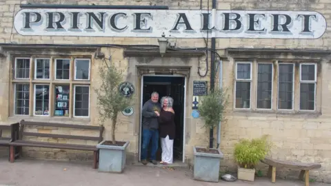 Lotte and Miles stand together in the doorway of their pub. They are holding hands and smiling underneath a large sign which reads 'Prince Albert'. 