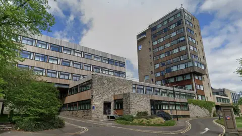 A grey/brown building at least 11 storeys high with windows. It is connected to a smaller two or three storey building of the same material on one side. On the other side connected to the smaller building is a taller building. A pathway leads up to the buildings.